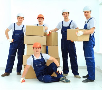 A group of five professional removal workers dressed in blue overalls and safety helmets inside a bright, modern residential space, likely a house or apartment in Marylebone, London. The workers are positioned around a stack of cardboard moving boxes of various sizes, with some holding or supporting the boxes, ready for transport. The background includes plain white walls and a light-colored wooden floor, creating a clean and organized environment for house removals. One worker is seated on the floor in front, smiling, with a cardboard box nearby, while the other four stand behind, engaged in their tasks, with some supporting or preparing to lift boxes. The scene captures the process of packing or preparing items for relocation, highlighting careful handling of household belongings such as boxes, possibly containing personal items, and the use of safety equipment like helmets for consideration during removals in the Marylebone area. The natural light emphasizes the professional yet approachable atmosphere of the removals team working efficiently within a typical residential interior in London.