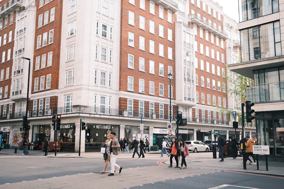 A busy street scene in Marylebone with a large multi-storey building featuring a mix of red brick and cream-colored façade, with multiple windows and a corner entrance. On the ground floor, there are
