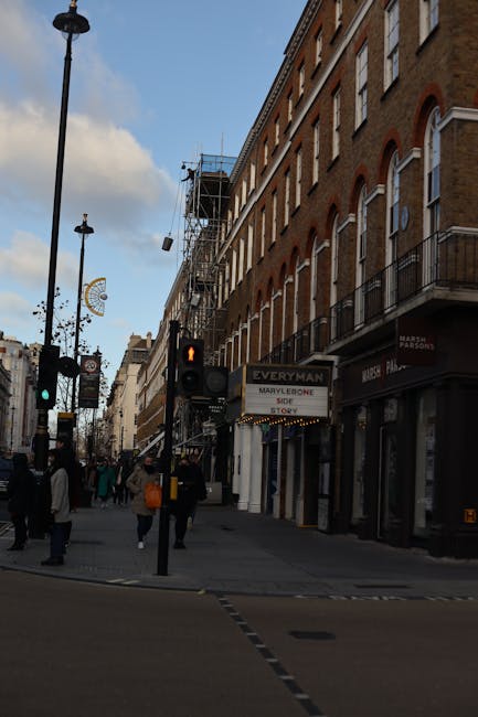 A busy street scene in Marylebone with a large multi-storey building featuring a mix of red brick and cream-colored façade, with multiple windows and a corner entrance. On the ground floor, there are